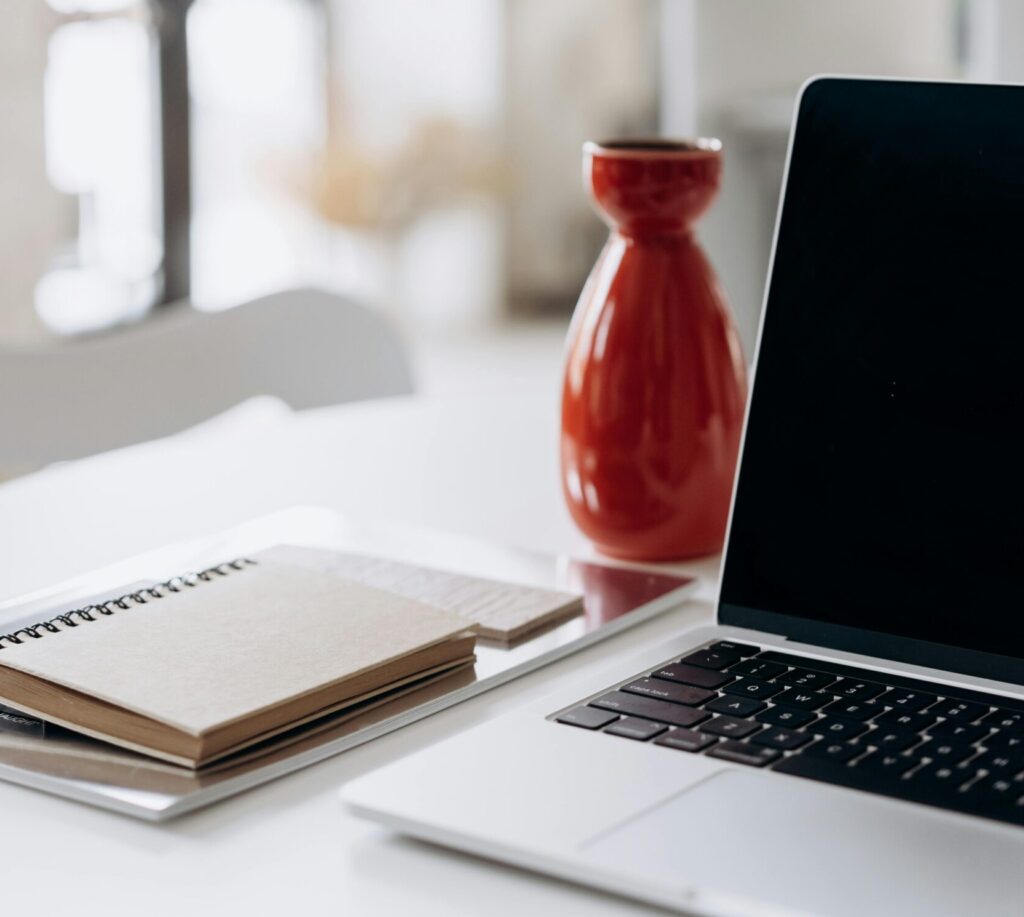 A minimalist home office setup featuring a laptop, notebook, and a red vase on a white desk.